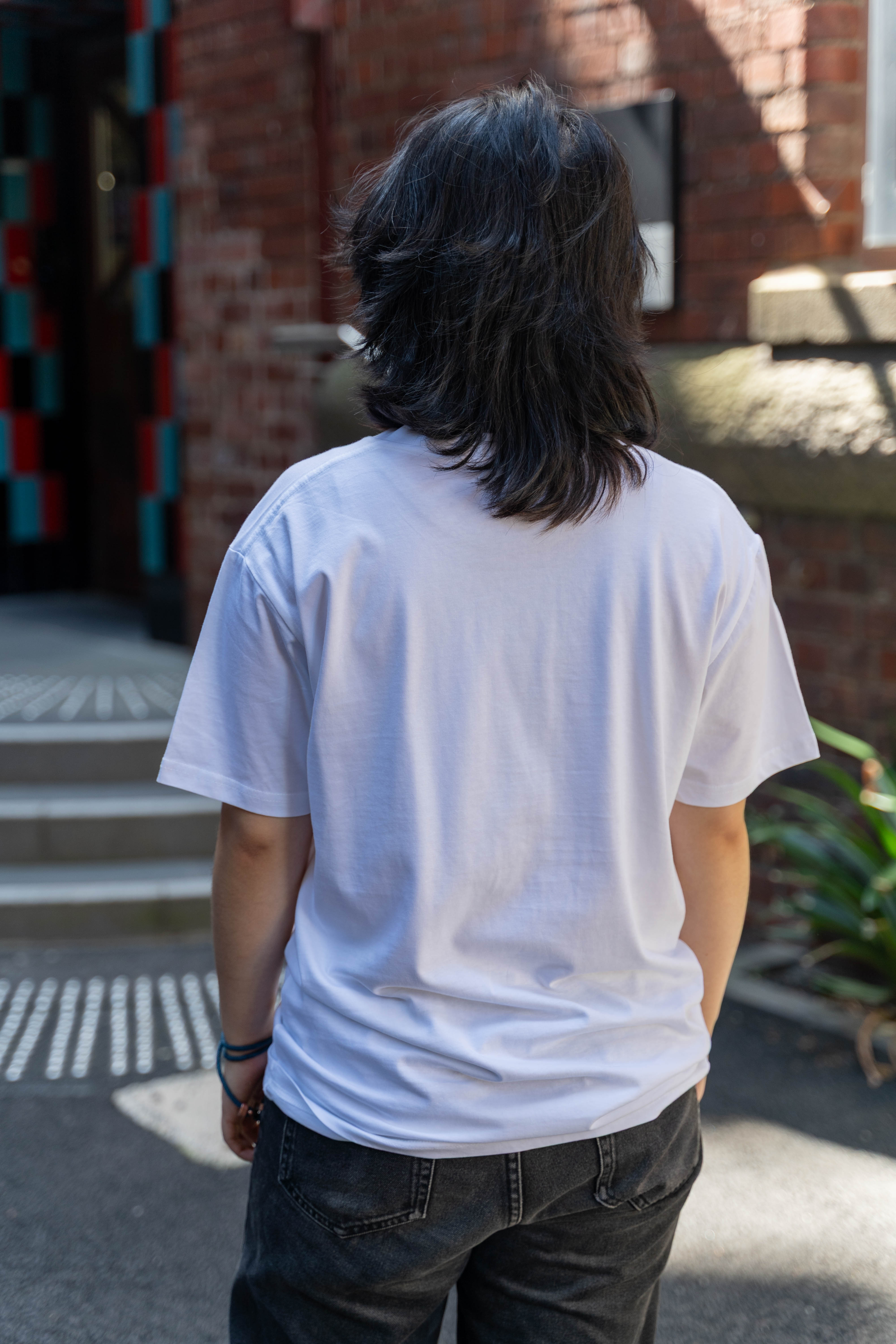 Model wears the Limited Edition Pride Unity Tee in white, presenting the plain back design while standing outdoors in front of a brick building. The model is styled with dark jeans for a casual look. Available at the RMIT Store.